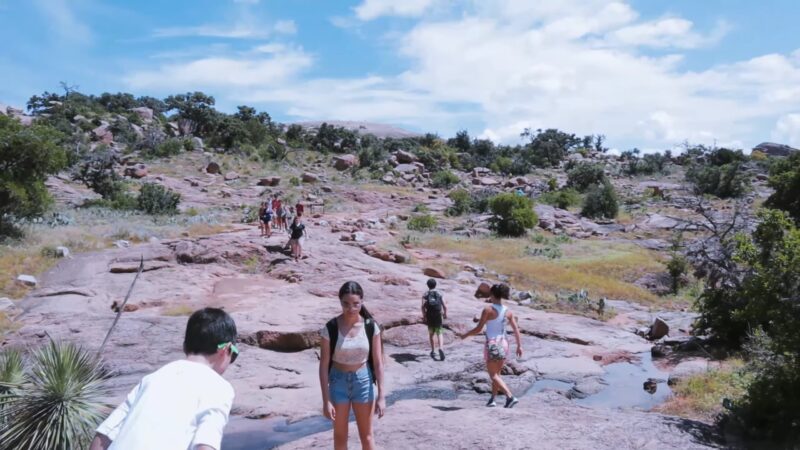 Hikers walking along the rocky Enchanted Rock Summit Trail, surrounded by vegetation and clear skies in Enchanted Rock State Natural Area